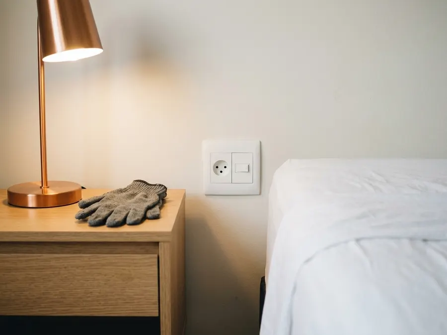 Tomada de três furos e interruptor ao lado da cama, com o abajur aceso